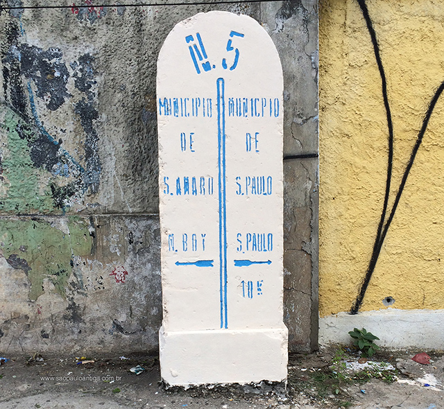 Foto do marco nº5 que dividia o município de São Paulo com o município de Santo Amaro. O marco é todo de pedra, pintado de branco e com sua parte escrita em letras de cor azul. A numeração do marco está na parte de cima, embaixo uma linha azul divide o marco no meio, em cada lado o nome de cada município, com uma seta logo abaixo apontando a direção de cada uma das cidades. Bem atrás do marco temos dois muros, um de cor cinza e o outro de cor amarela. O marco está posicionado em uma calçada.