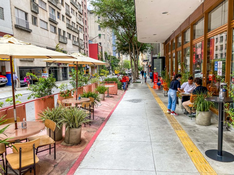 Do lado esquerdo há mesas e cadeiras pintadas de rosa, no meio a calçada, e do lado direito a fachada do estabelecimento