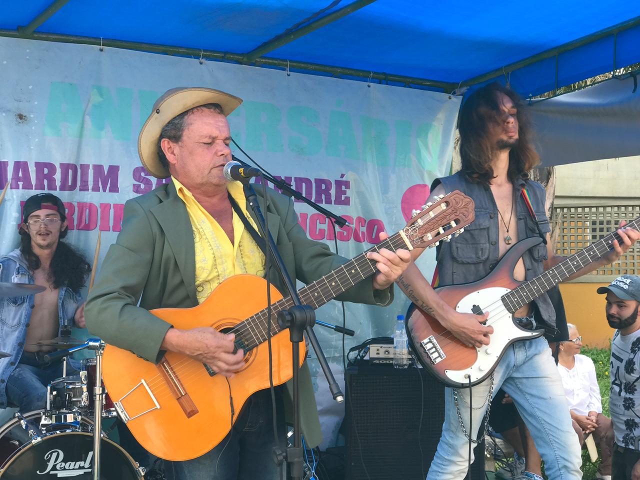 banda anima a festa de aniversário do Jardim Santo André e Jardim São Francisco. No palco, em primeiro plano, homem de chapéu, camisa amarela, aberta em dois botões, blazer verde escuro, calça jeans, toca violão elétrico, de olhos fechados. Ao lado, guitarrista jovem, sem camisa, colete aberto, cabeludo, de cavanhaque, toca o instrumento. Atrás da placa de aniversário, grande, está o baterista, jovem, cabeludo, também sem camisa e um colete jeans, de óculos.
