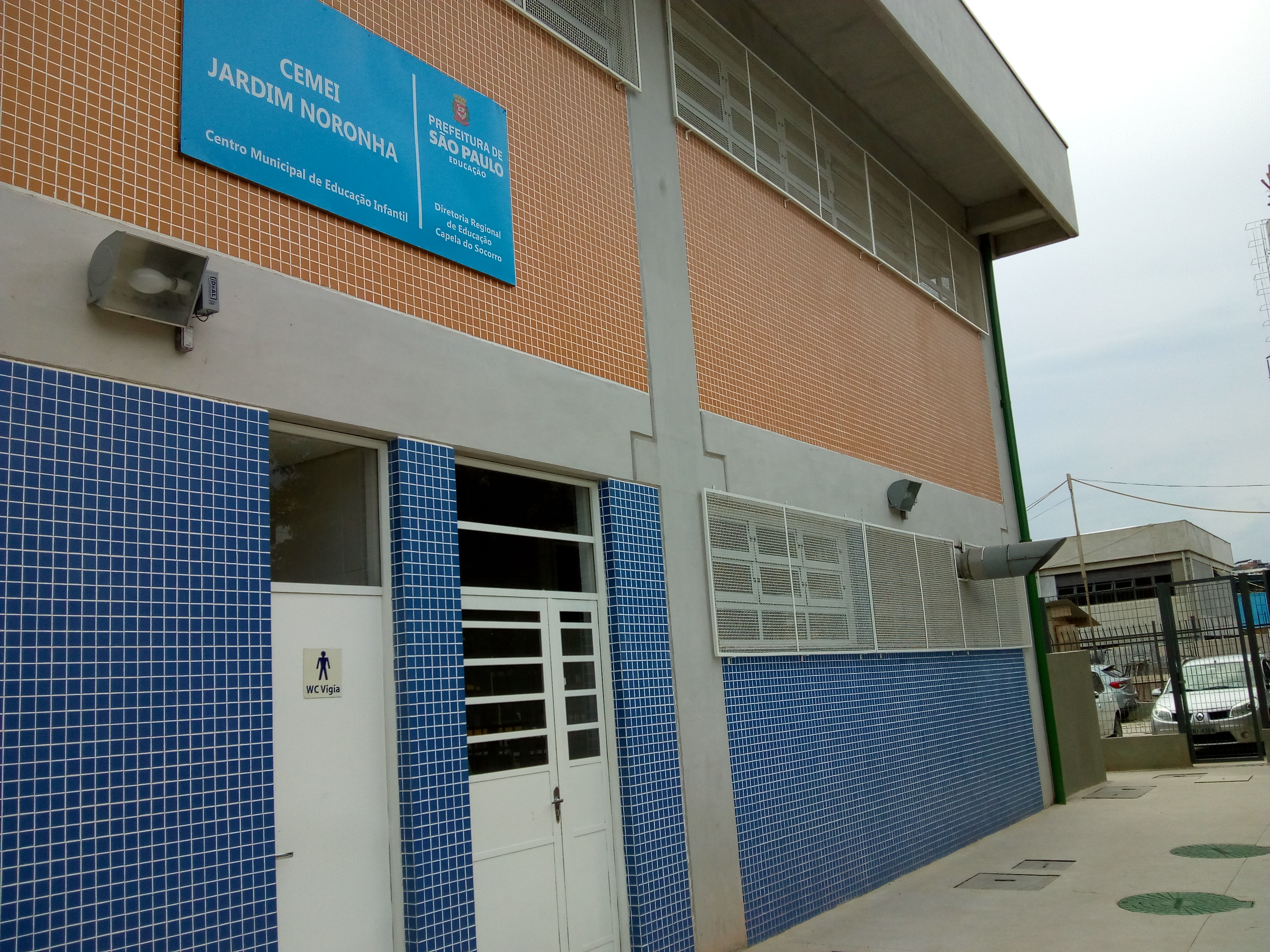 #PraCegoVer - Fachada do CEMEI Jardim Noronha. A unidade é construída em colunas e vigas de concreto aparente. Os intervalos são cobertos de pastilhas azuis e laranja.