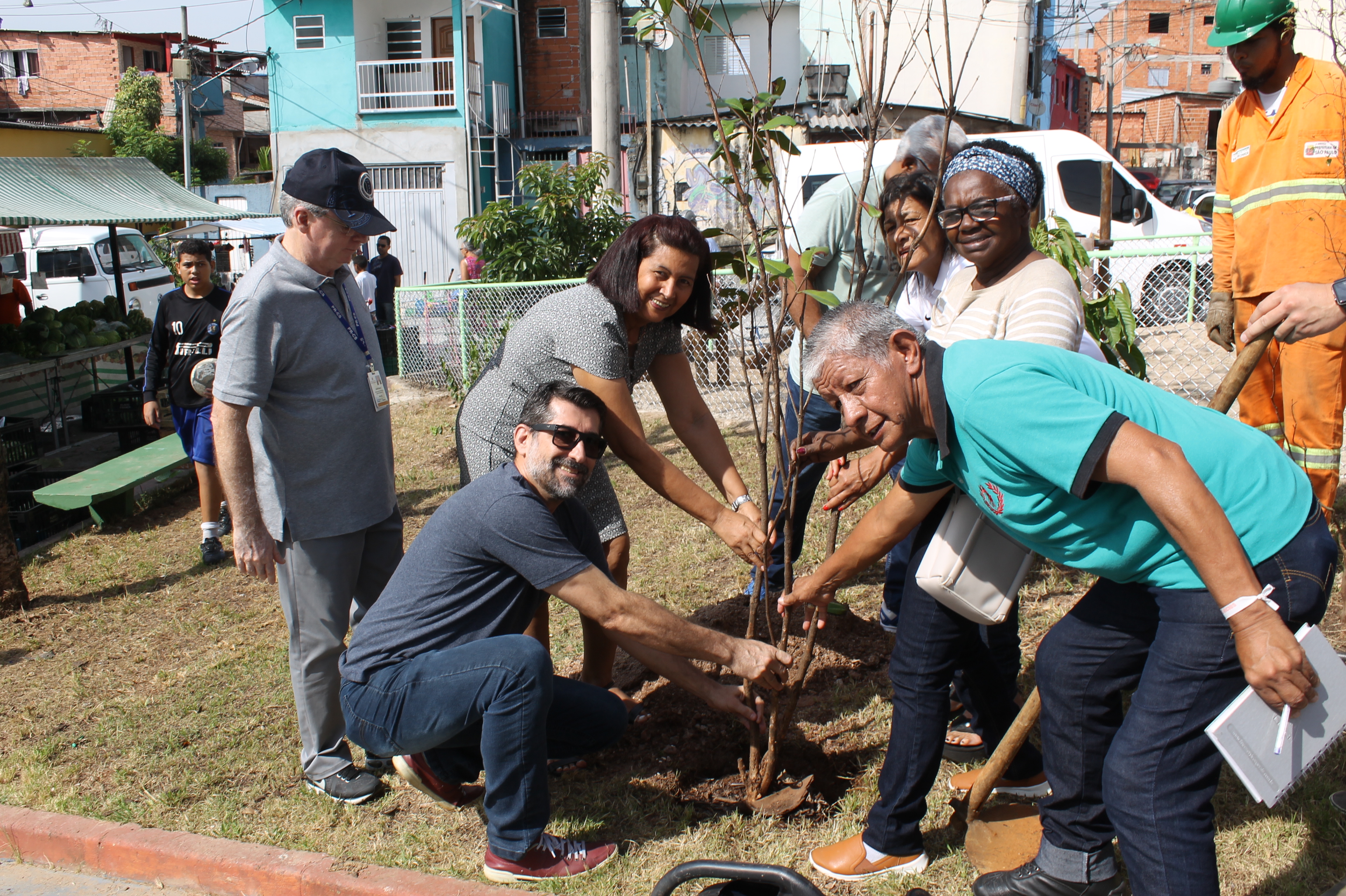 #PraCegoVer - Pessoas plantam muda de árvore na praça da Vila da Paz