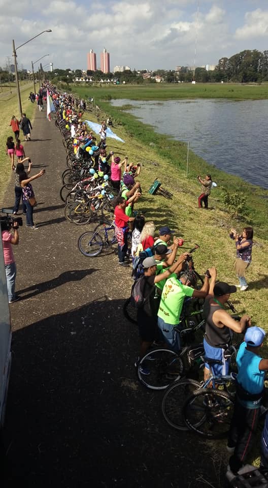 #PraCegoVer - Centenas de pessoas à beira da represa Guarapiranga, no dia do Abraço à represa. Ciclistas, pessoas a pé, fotógrafos, com a água quase batendo nos pés.