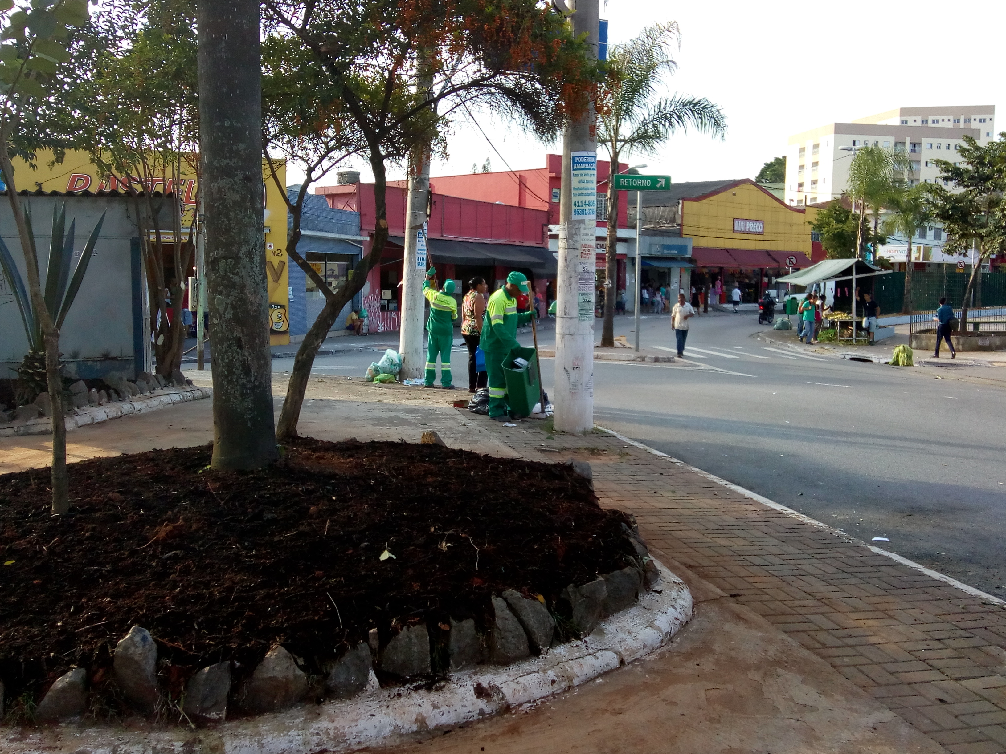 #PraCegoVer - No primeiro plano, um canteiro com uma árvore e mudas de flores. Trabalhadores da Prefeitura, em macacões verde-amarelos, limpam a praça. Ao fundo, as casas comerciais da região