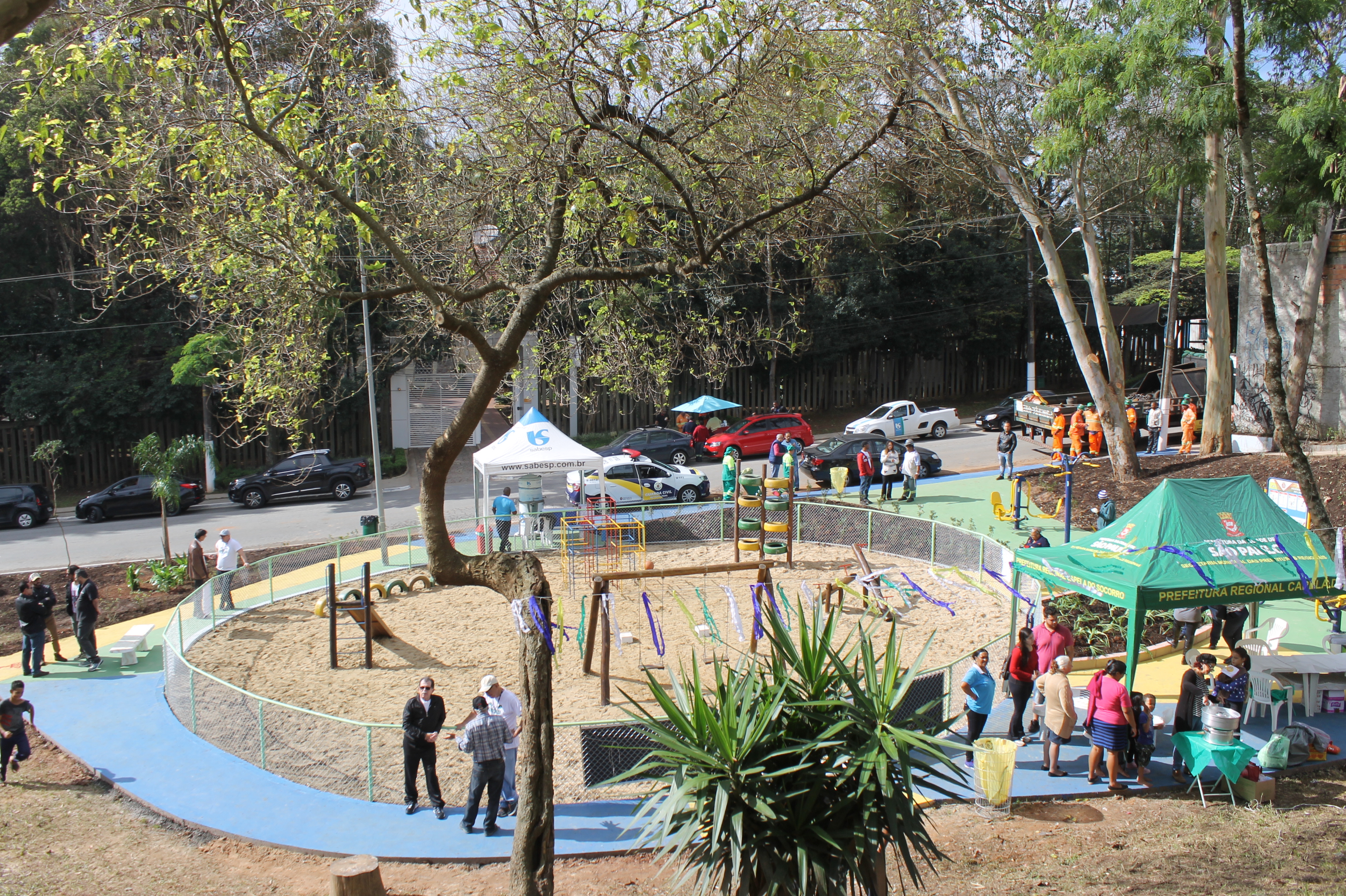 #PraCegoVer – No espaço central, redondo, da praça, aparecem brinquedos como escorregador, balanços e trepa-trepa. Do lado direito, uma tenda verde, com a inscrição Prefeitura Capela do Socorro. Ao fundo, outra tenda, branca, da Sabesp. Trabalhadores e visitantes espalham-se pela praça, bem em frente ao portão de entrada do Senac, no Jardim dos Bichinhos.