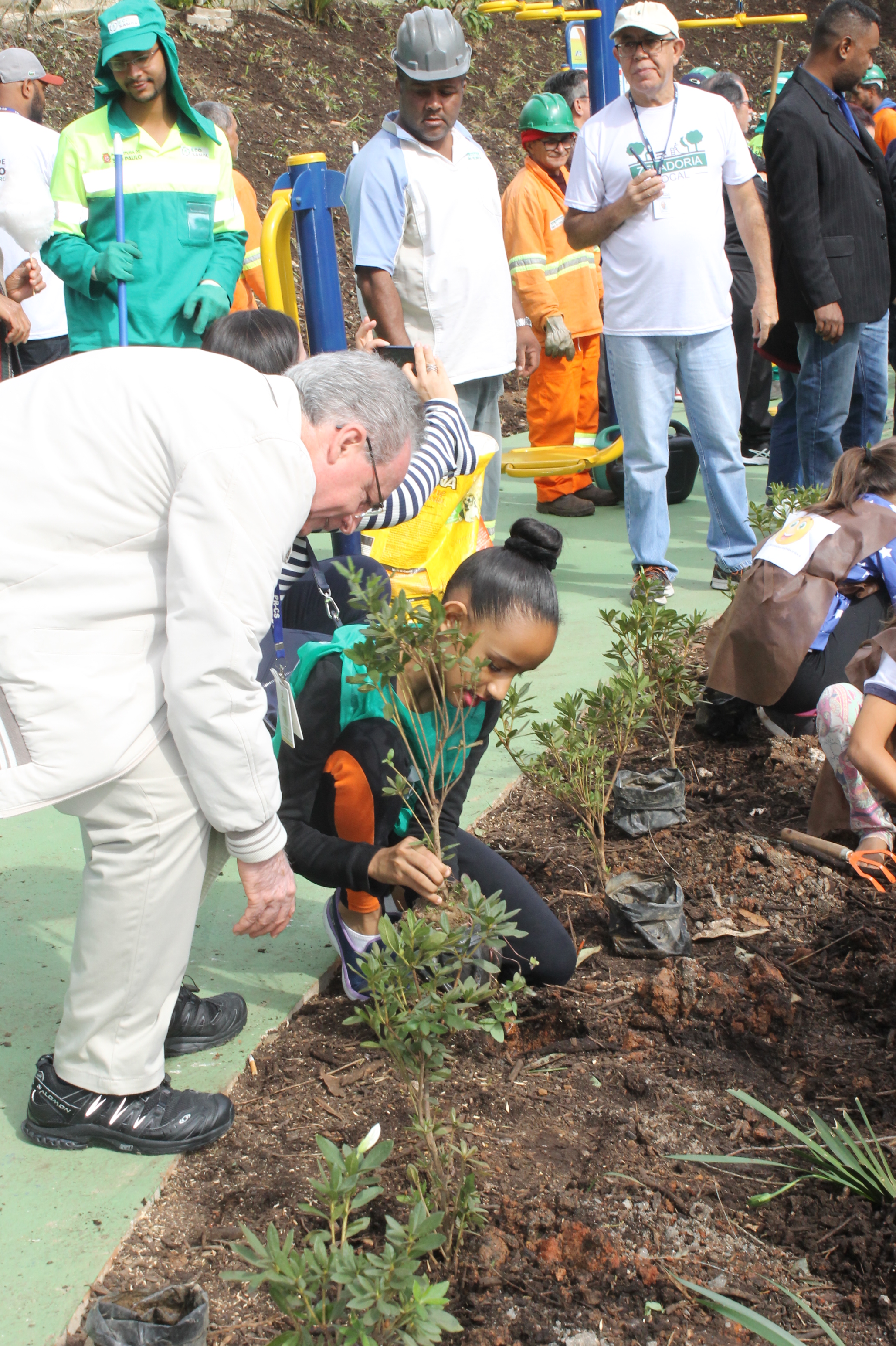 #PraCegoVer – Há um canteiro do lado direito, embaixo, da foto, com crianças plantando mudas. Do lado esquerdo, uma menina de uniforme azul-marinho e verde é ajudada pelo subprefeito João Batista de Santiago.