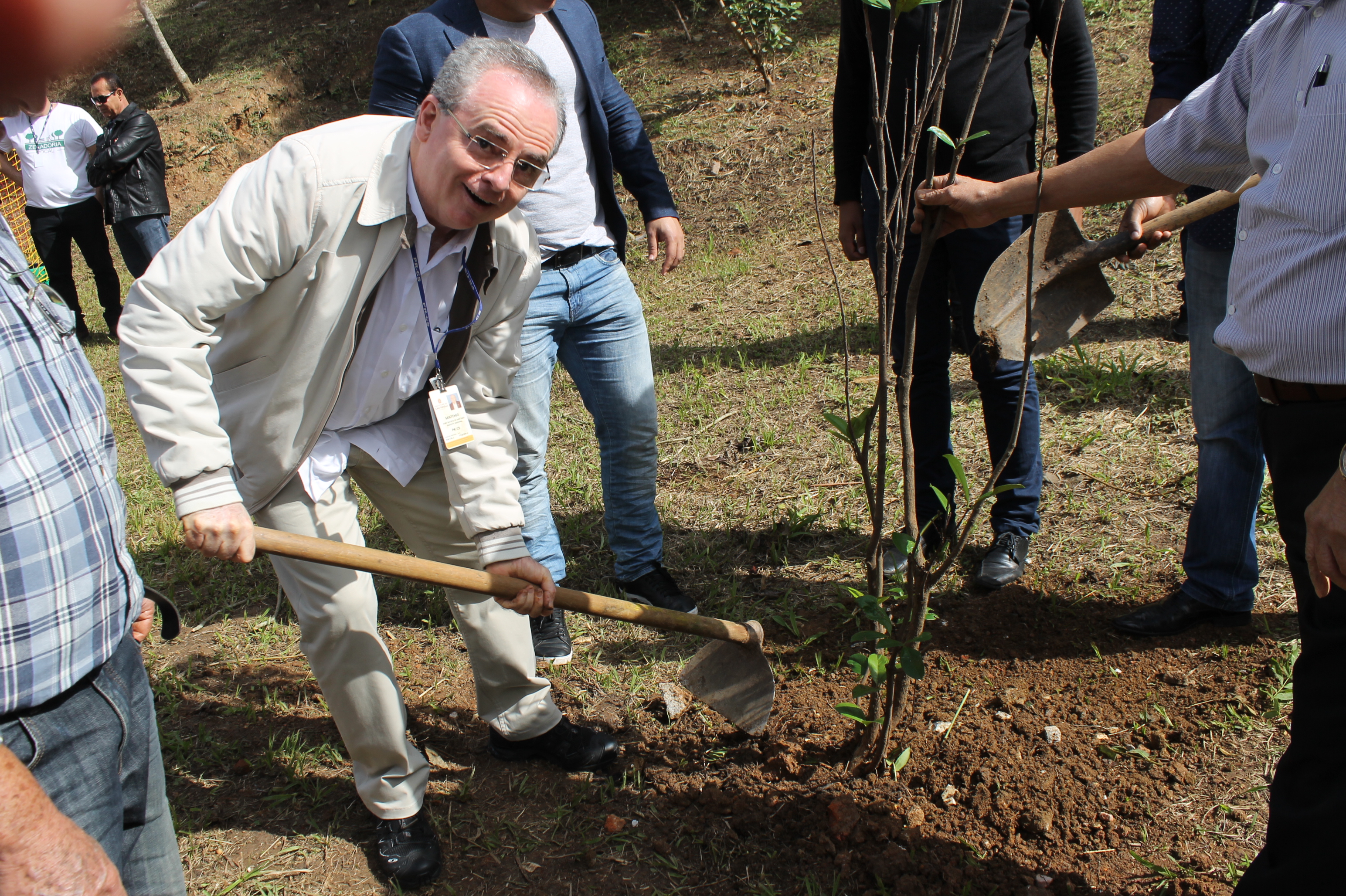 #PraCegoVer – O subprefeito João Batista de Santiago maneja uma enxada para plantar muda de árvore. Um munícipe segura o tronco da muda para mantê-la em pé.