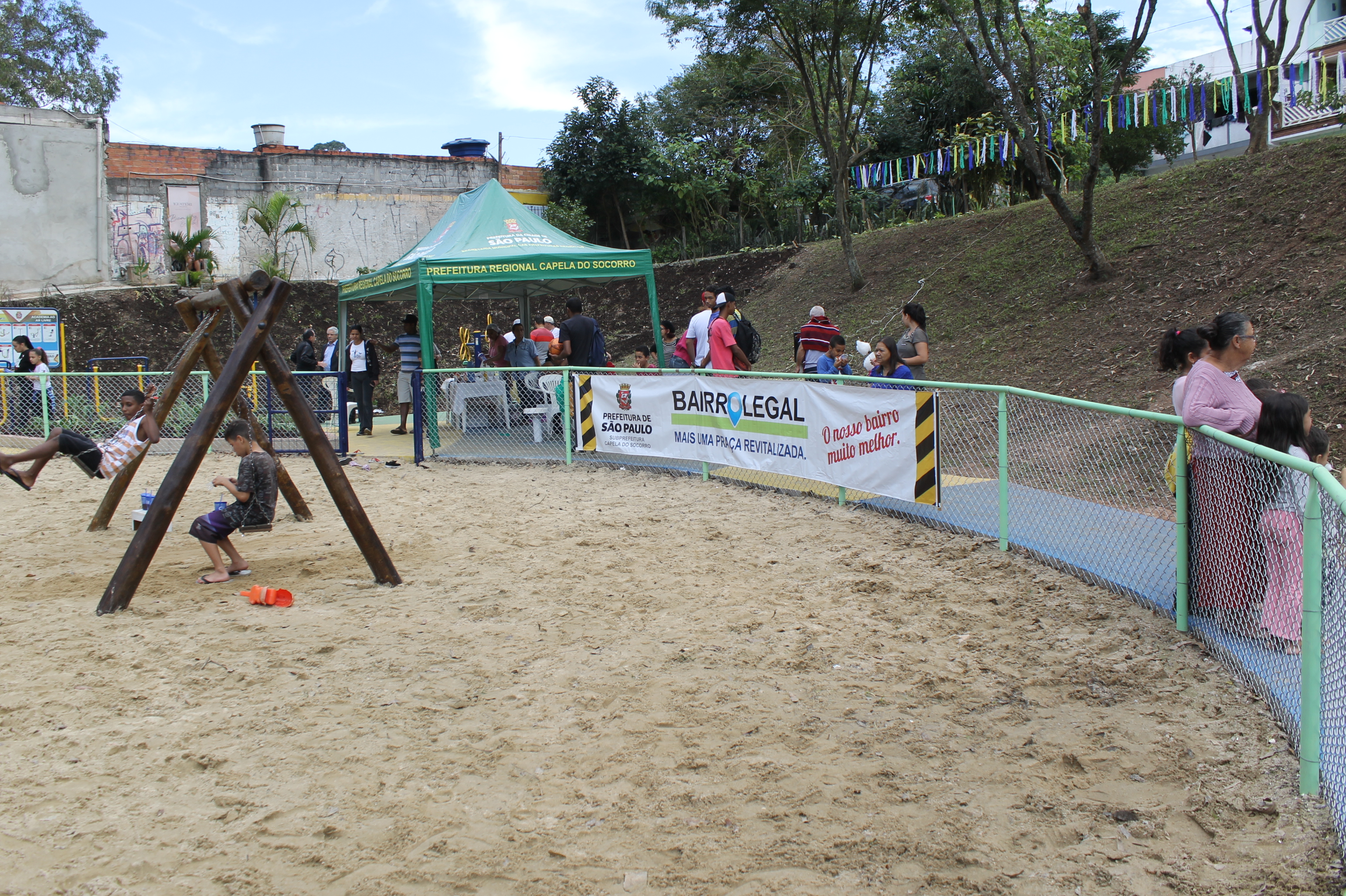 #PraCegoVer – Caixa de areia, cercada de tela, para proteger as crianças brincando. À esquerda, dois meninos brincam no balanço. Uma faixa, na área central, avisa: 