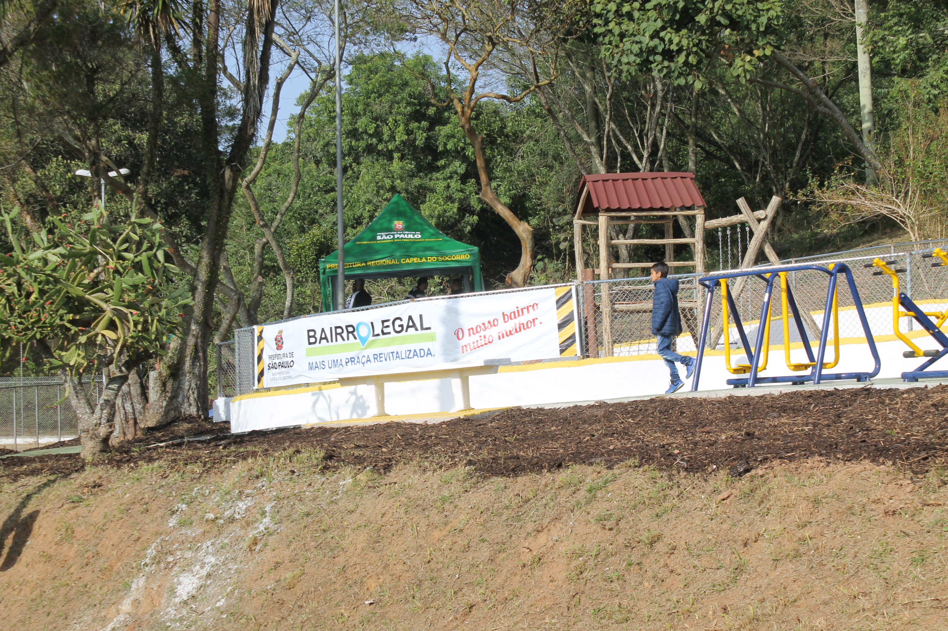 #PraCegoVer - Caixa de areia para crianças na praça da segunda Balsa na Ilha do Bororé. Há uma faixa anunciando a inauguração do local. Do lado direito, aparelhos de ginástica. Um menino passa ao lado do alambrado. Bem no fundo, há um gazebo da Subprefeitura.