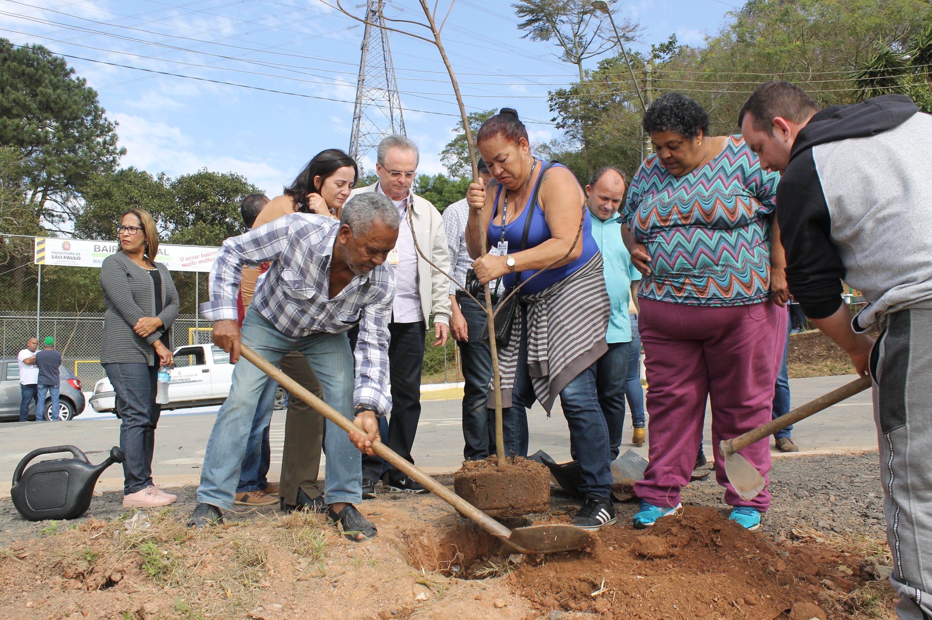 #PraCegoVer - Moradores do Jardim Santa Tereza plantam muda de árvore. Um senhor coloca terra no buraco com uma pá. Uma senhora sustenta a muda.