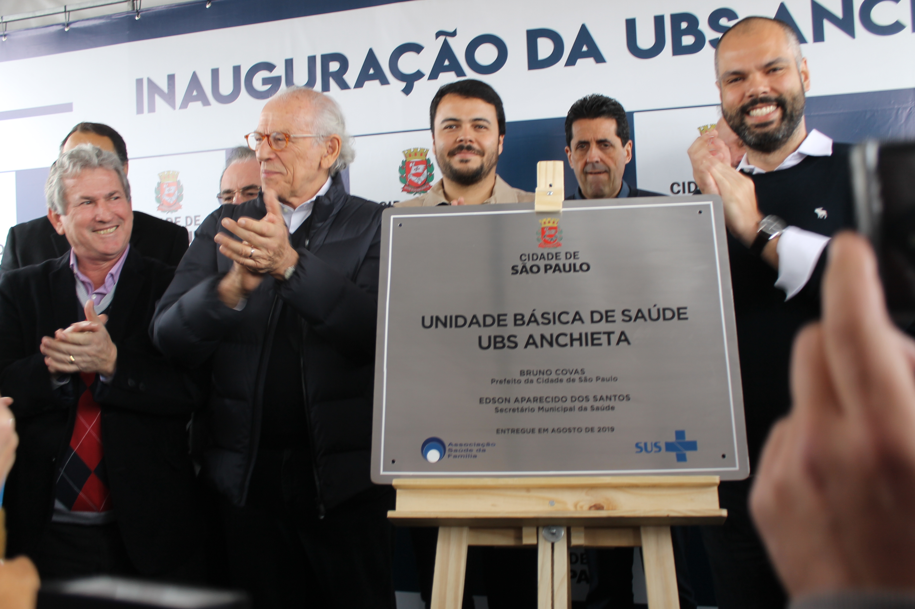 #PraCegoVer - Prefeito Bruno Covas descerra a placa da inauguração da UBS. Todos aplaudem. Na placa, se lê: Cidade de SãoPaulo - Unidade Básica de Saúde - UBS Anchieta - Bruno Covas, prefeito da Cidade de São Paulo - Edson Aparecido dos Santos, secretário municipal de Saude - Entregue em agosto de 2019 - De um lado o logotipo da Associação Saúde da Família (ASF) e, de outro, o logotipo do SUS.