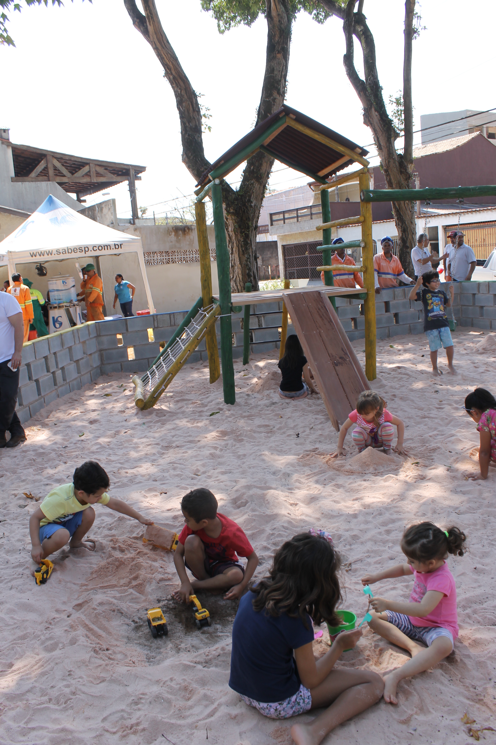 #PraCegoVer - Crianças brincam na caixa de areia da praça. O local é cercado por um muro baixo de blocos. No centro da caixa, há um brinquedo chamado Casa do Tarzan, com escada de cordas e escorregador.