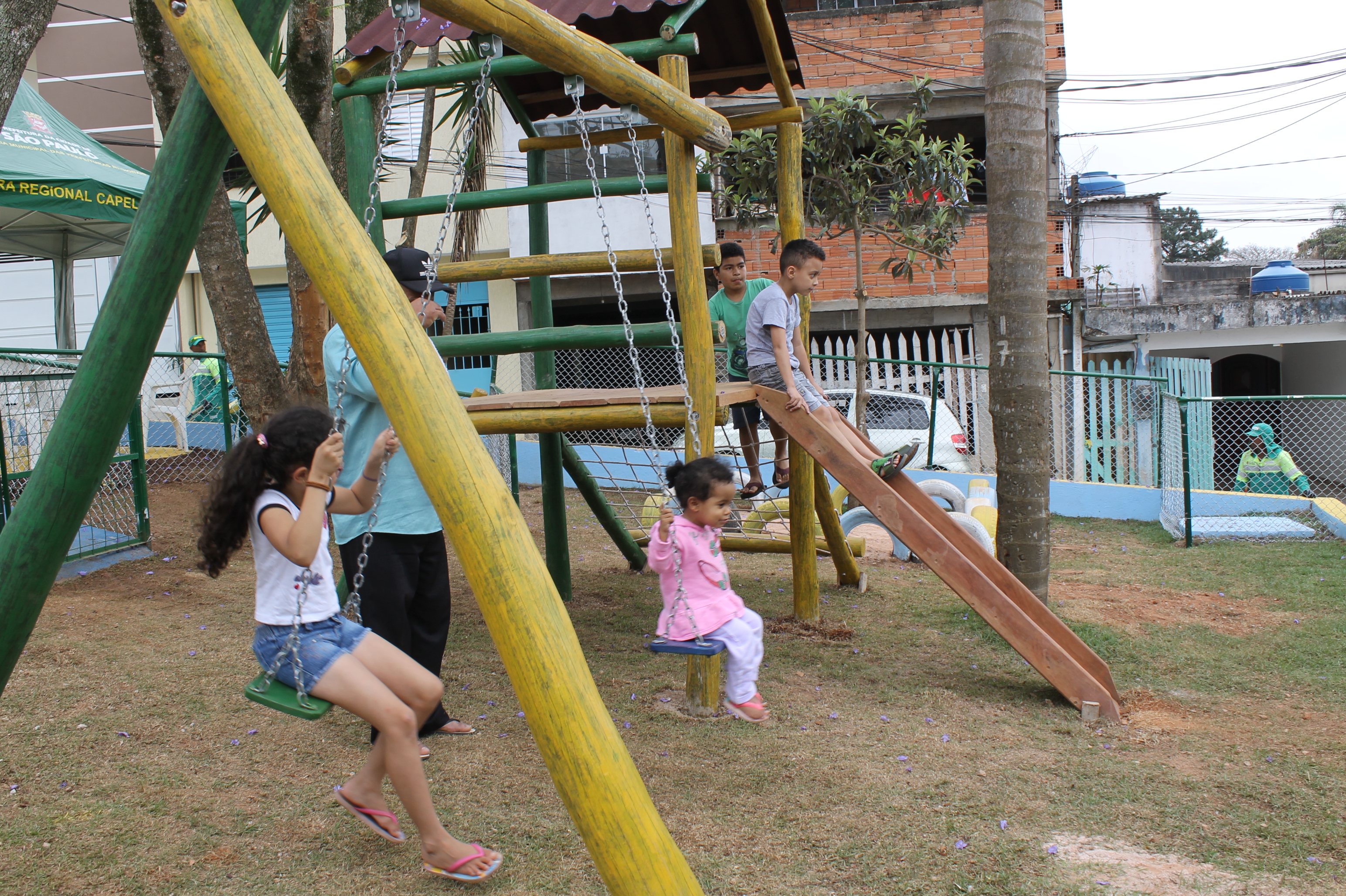 #PraCegoVer - Crianças brincam no balanço da praça. Um menino está no topo do escorregador. Ao fundo, as casas do local.