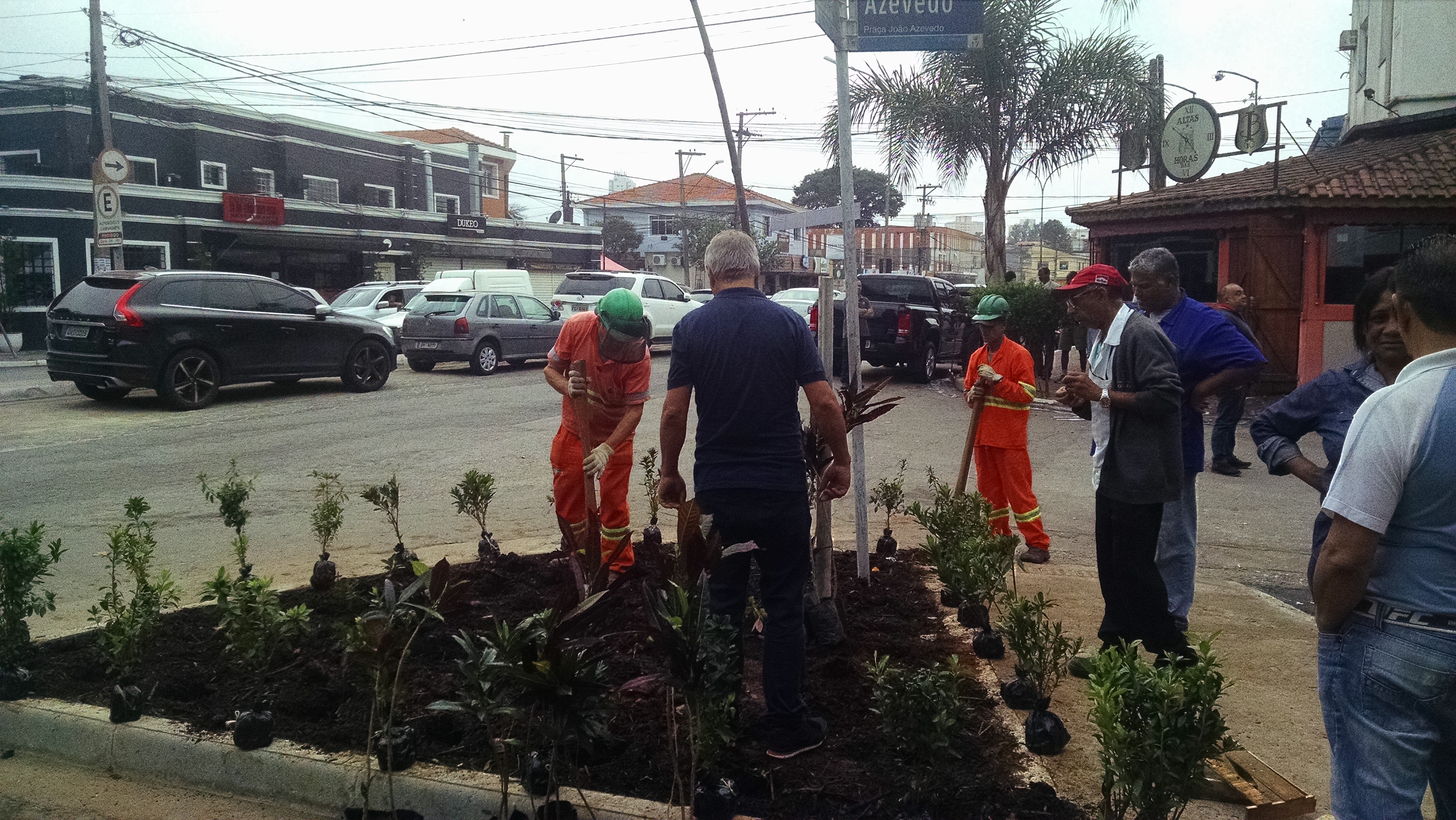 #PraCegoVer - Trabalhadores da Subprefeitura e moradores plantam mudas de flores em um canteiro de formato triangular. Ao fundo, carros estacionados na avenida Inácio Cunha Leme.