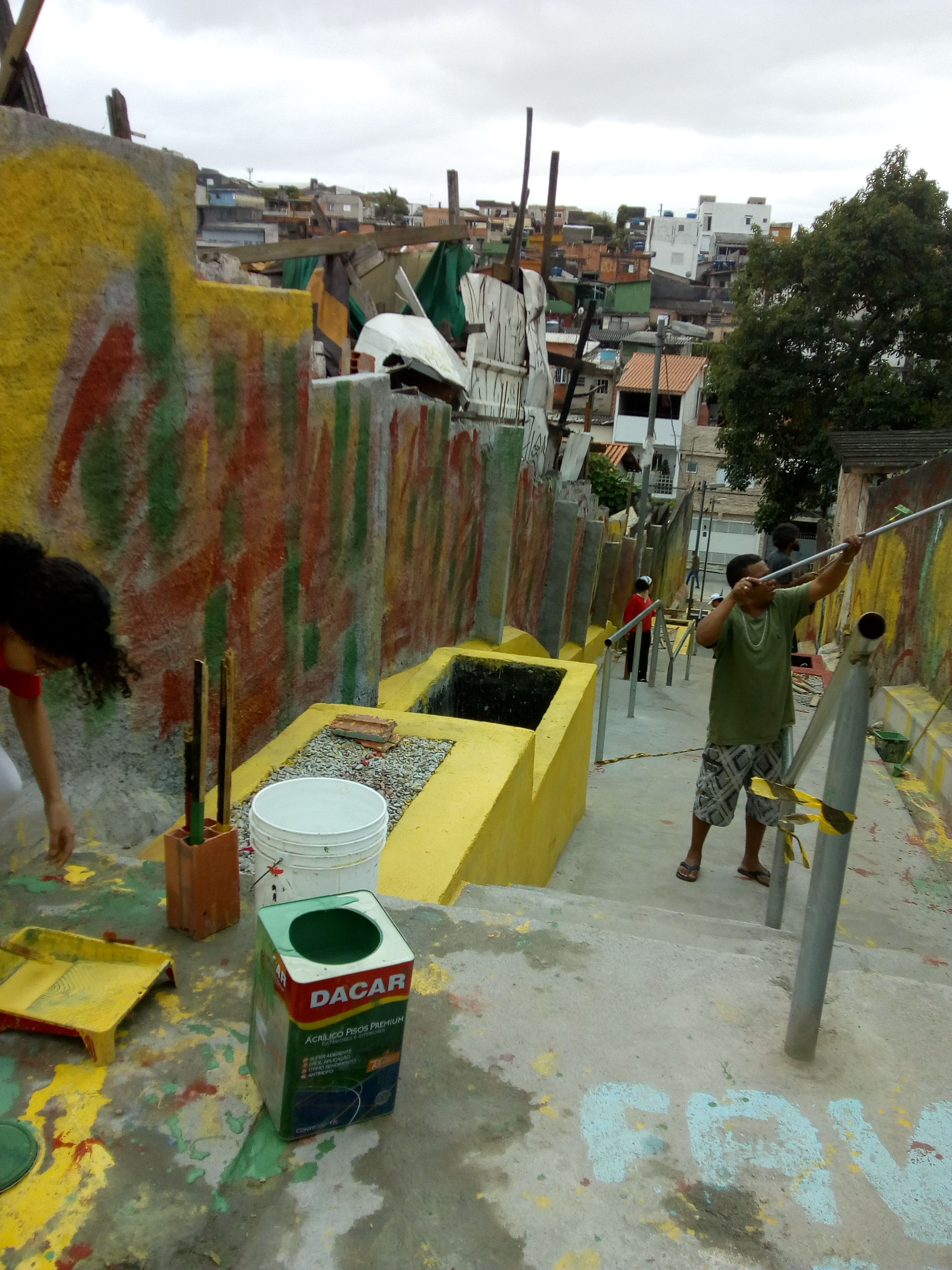 #PraCegoVer - Voluntários pintam os muros do escadão. As caixas de retenção de água da chuva estão pintadas de amarelo ou vermelho. No primeiro plano, há uma lata grande de tinta.