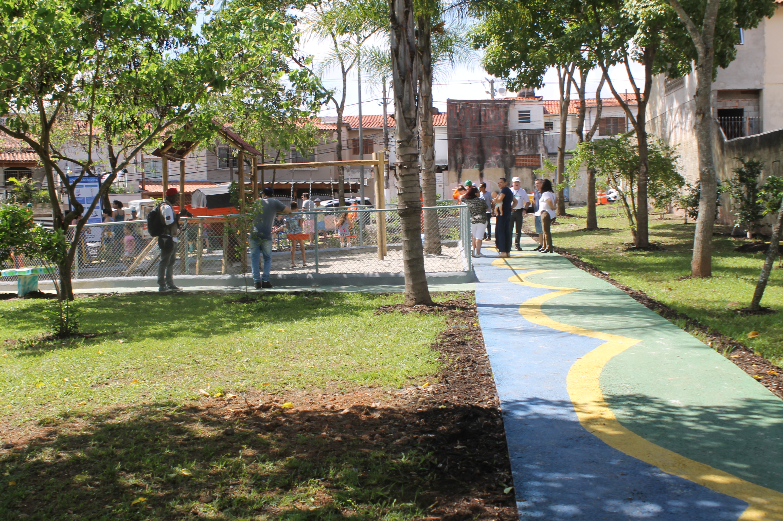 #PraCegoVer - Calçada que vai do primeiro plano ao fundo, com desenhos em azul, amarelo e verde. Do lado esquerdo, o ParCão — para exercitar animais de estimação. Árvores ponteiam a praça. Grupos de pessoas conversam do lado direito.