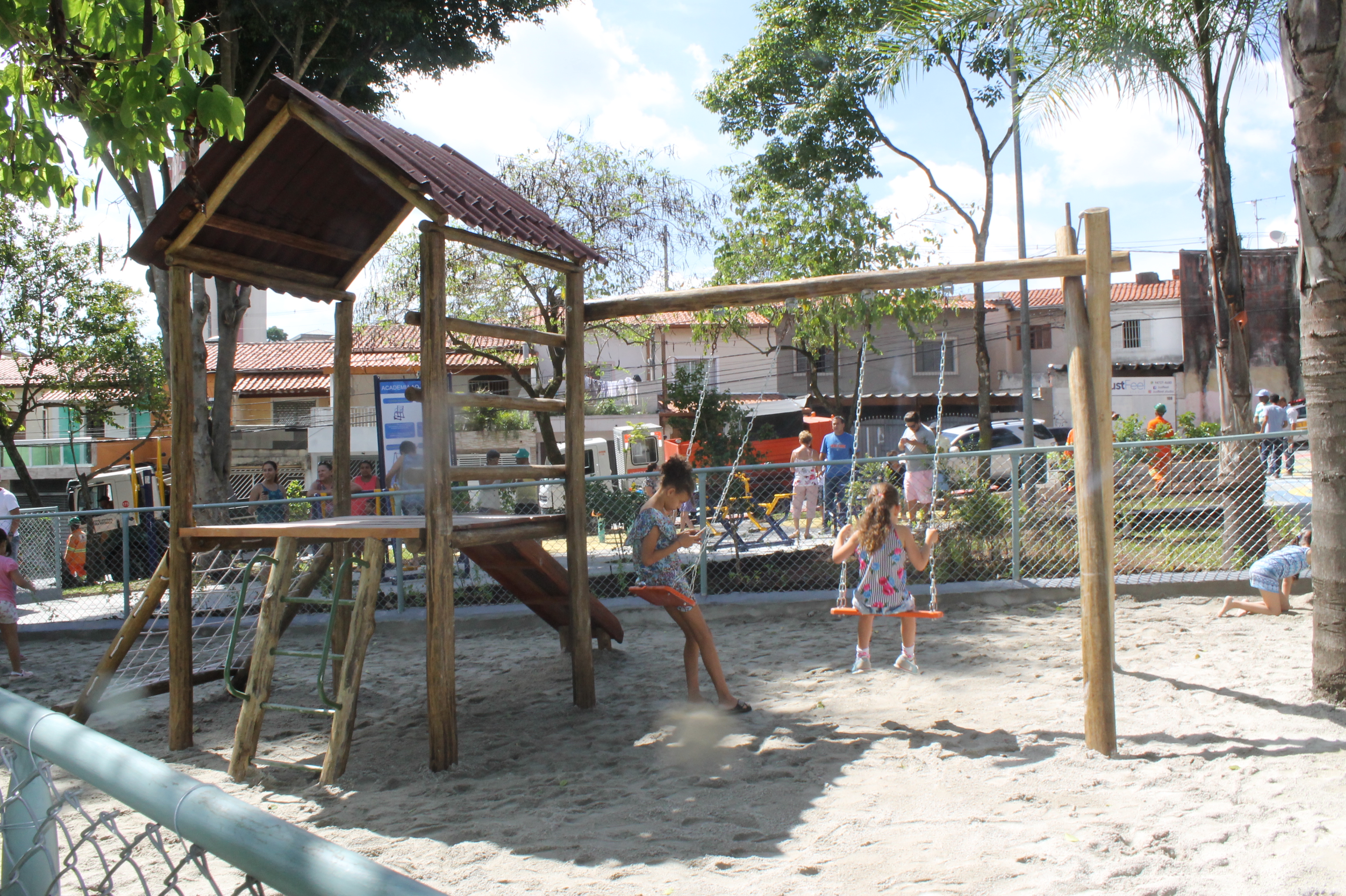 #PraCegoVer - Caixa de areia para crianças. Do lado esquerdo, a Casa do Tarzan, coberta, e com escada de madeira e rede de cordas, além de um escorregador. No meio, balanços, onde brincam duas meninas.