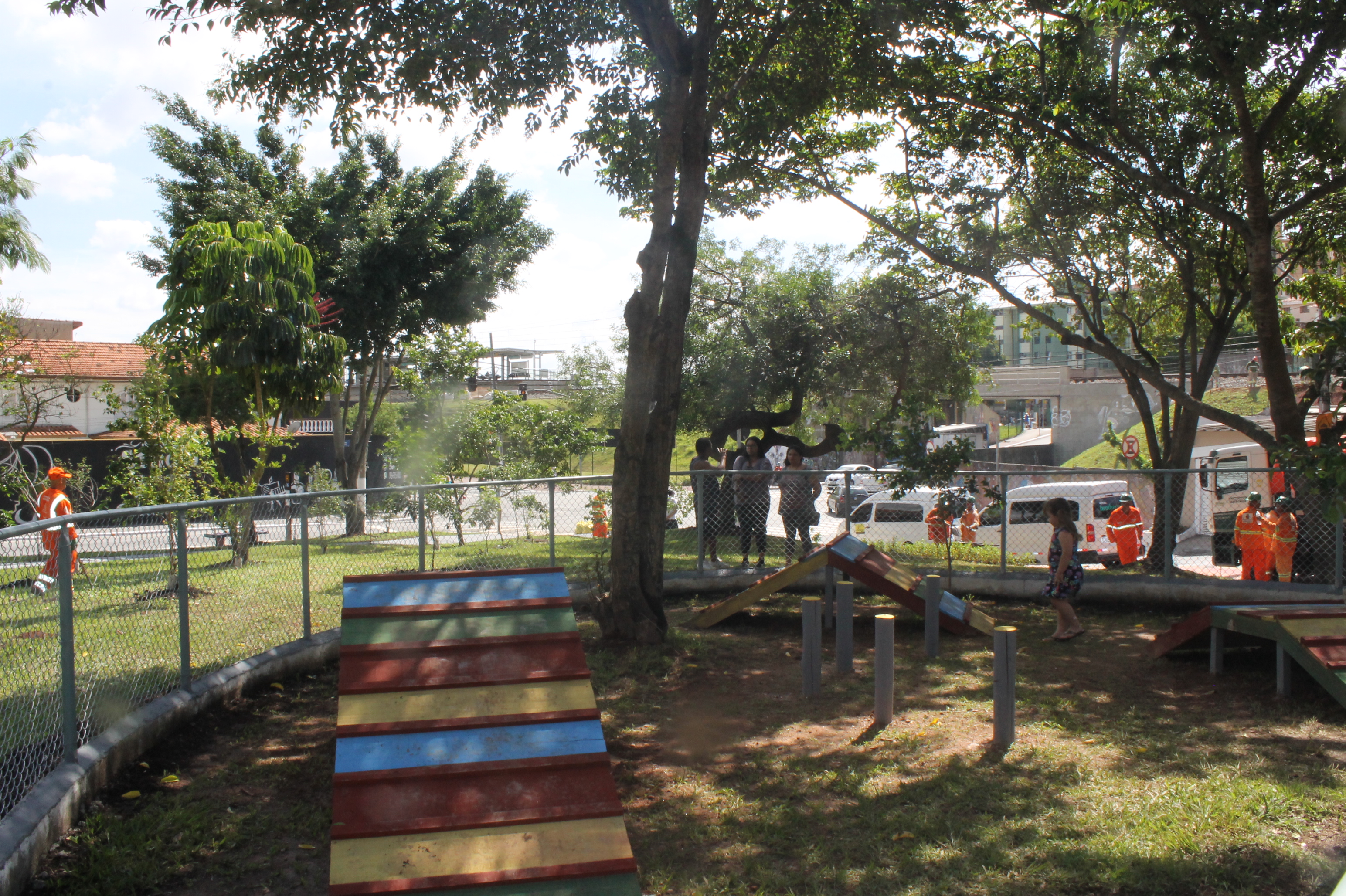 #PraCegoVer - ParCão para exercitar animais de estimação. A área, cercada, sob árvores, comporta três aparelhos de madeira, coloridas.