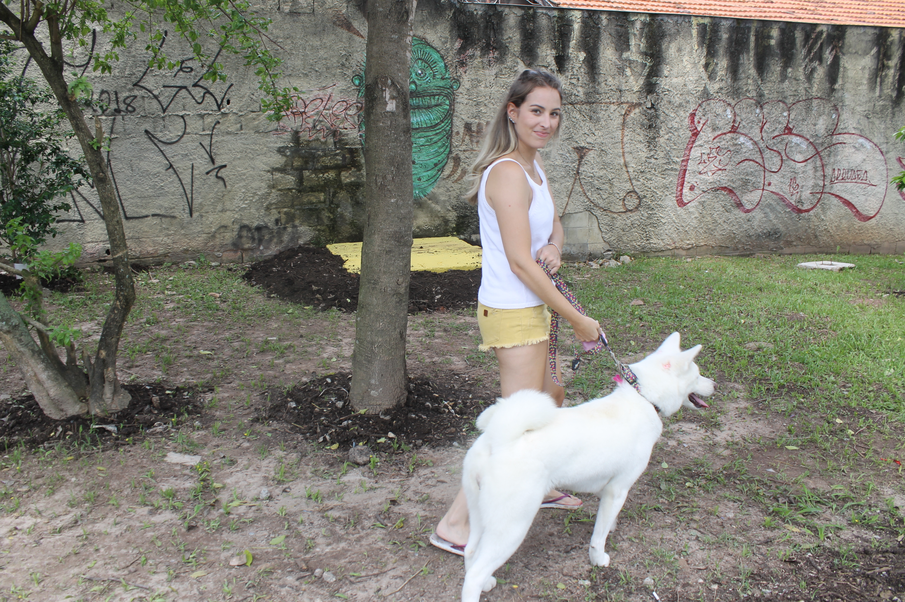 #PraCegoVer - Moradora da vizinhança com sua cadela toda branca. Ao fundo, árvores e um muro.