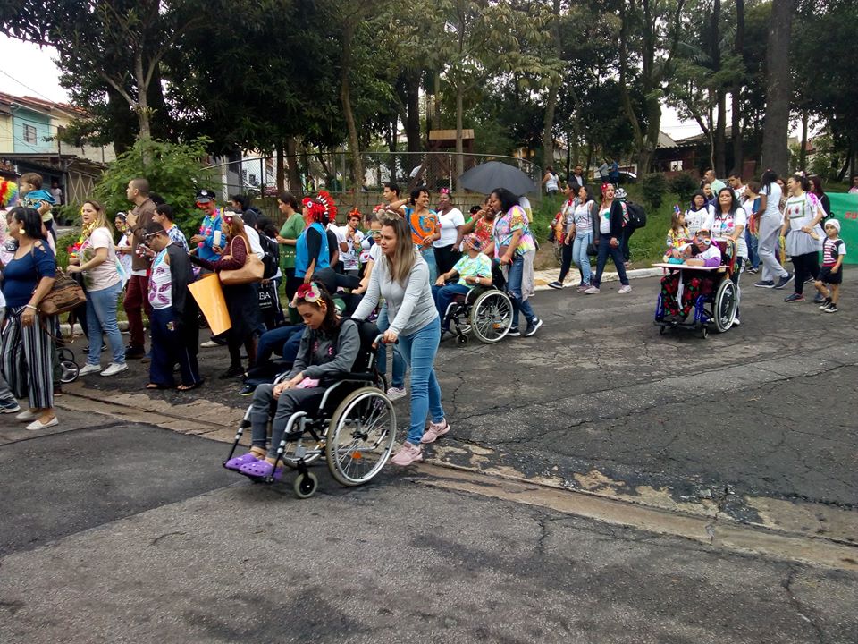 #PraCegoVer - Pessoas acompanham o desfile do bloco Integra Folia. Pode-se ver vários foliões conduzidos em cadeiras de rodas. Ao fundo, a praça Adriano Emídio de Sousa, no Jardim Clipper.