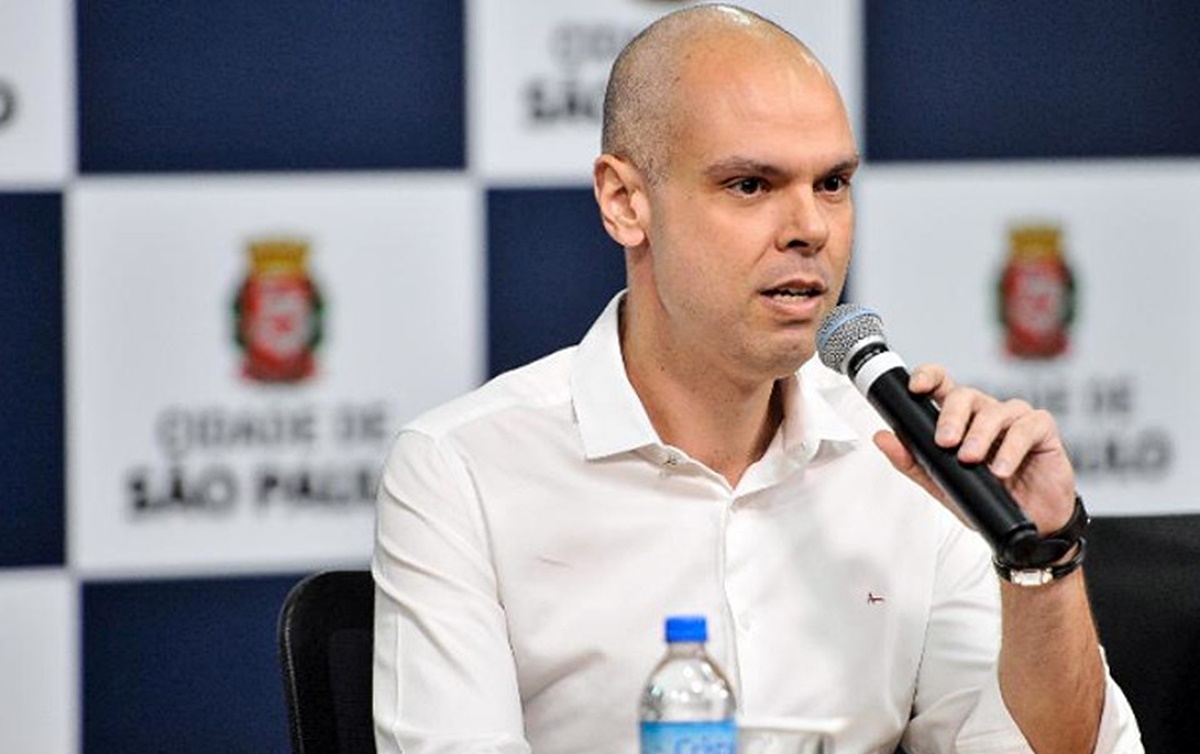 ParaCegoVer - O prefeito paulistano Bruno Covas anuncia medidas para enfrentar o coronavírus. Ele está de camisa branca contra um fundo com brasões do municípios.