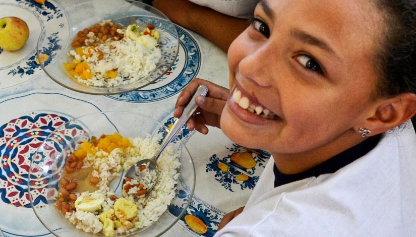#PraCegoVer - Criança sorrindo e olhando para a câmera, com prato de comida à sua frente.