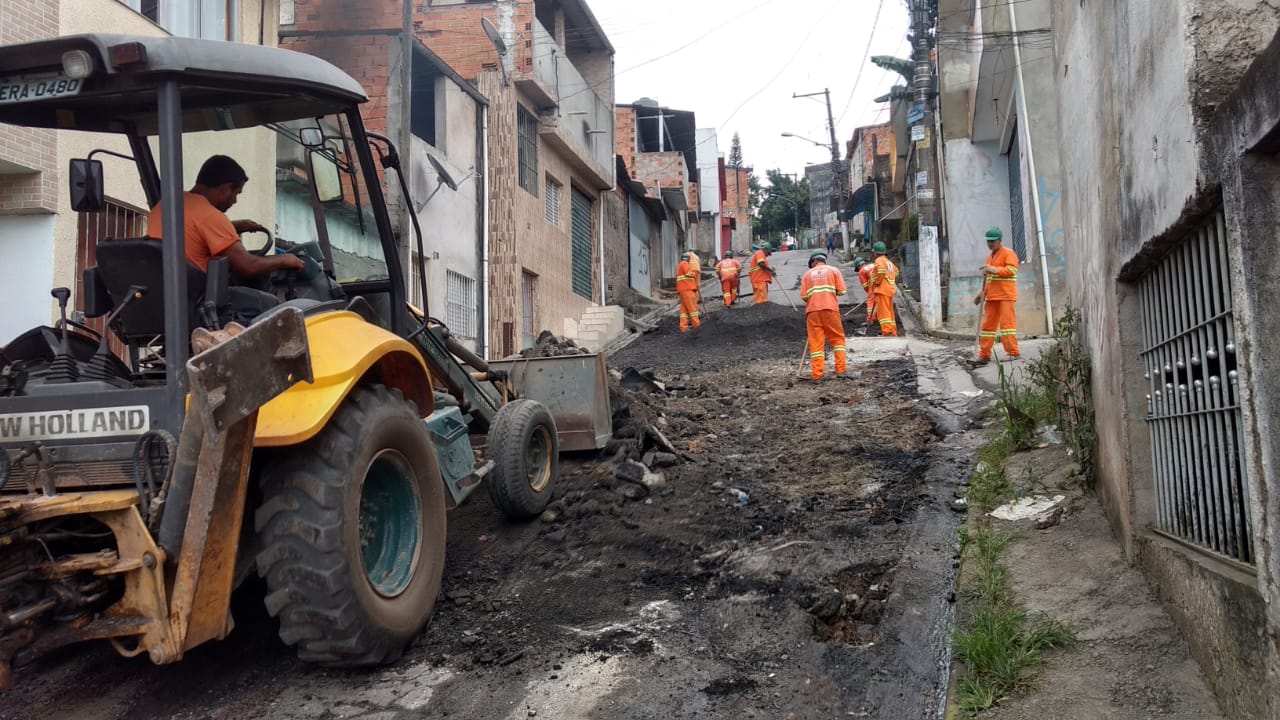 #PraCegoVer - Leito da rua em processo de nivelamento pela escavadeira. Outros trabalhadores espalham pó de asfalto para fixação da base.