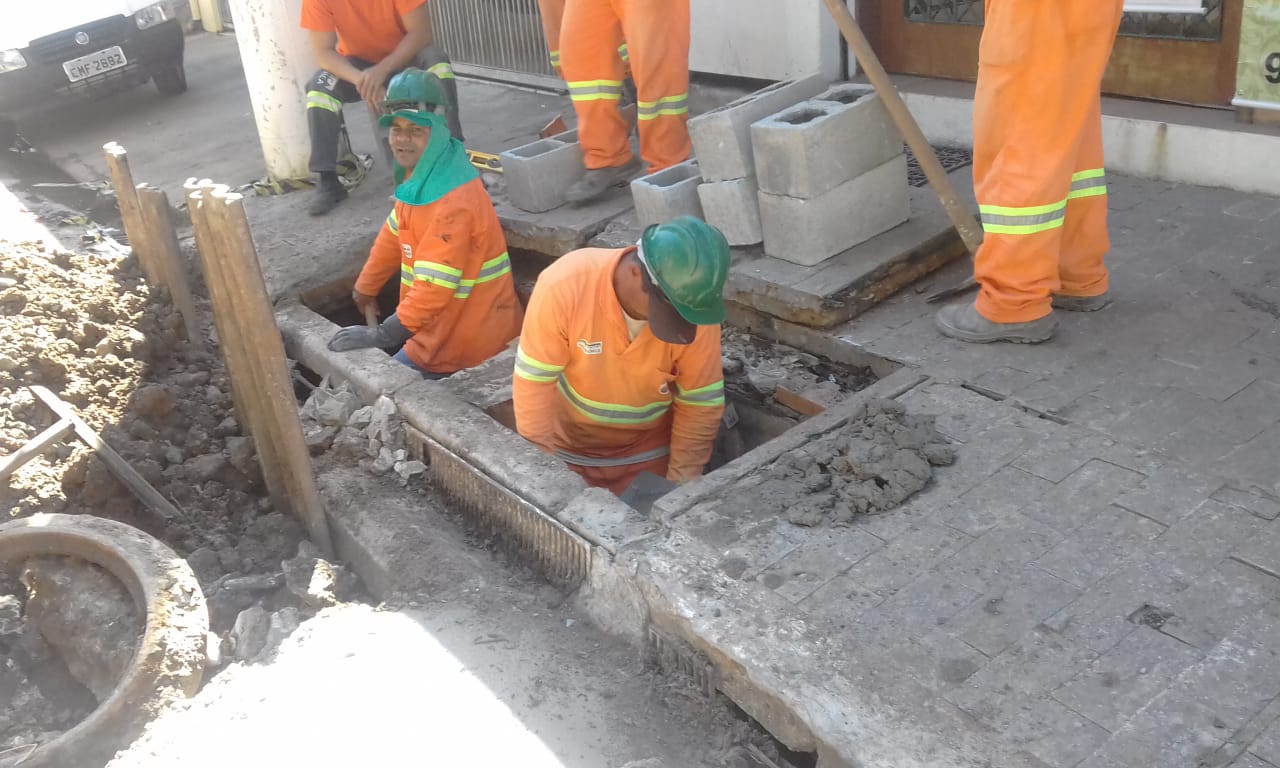 #PraCegoVer - Trabalhadores em macacões laranja e branco reformando boca-de-lobo (BL). Dois homens estão dentro da boca-de-lobo. Há restos de escavação no meio-fio. Na calçada, blocos de cimento.