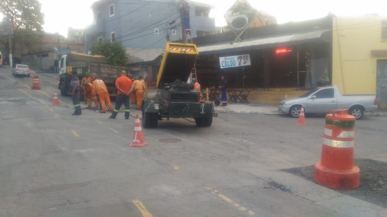 #PraCegoVer - Trecho da avenida com homens, caminhão e aparelhamento para tapar buracos. A via está marcada com cones laranja e branco. O buraco a ser consertado está ainda aberto.