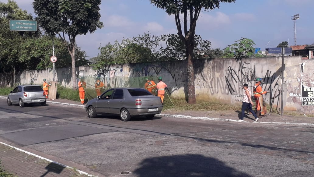 #PraCegoVer - Trabalhadores cortam grama na rua Miguel de Barros. Dois dos trabalhadores seguram proteção de tela para evitar o espalhamento da grama na via. Dois carros estão passando no momento.
