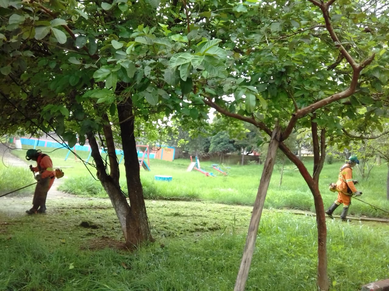 #PraCegoVer - Dois trabalhadores, um em cada lado da foto, cortam grama na via. Em primeiro plano, veem-se árvores. Ao fundo, brinquedos de playground.