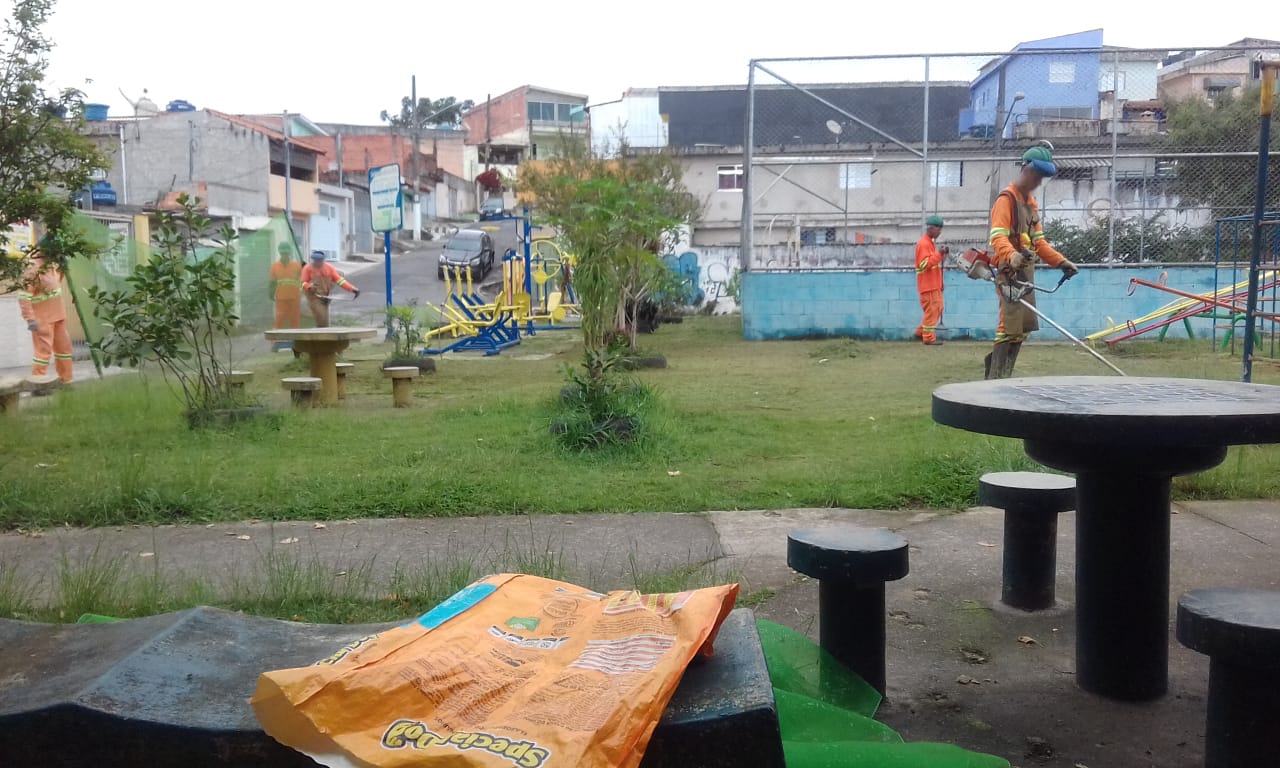 #PraCegoVer - Trabalhadores cortam grama em um espaço com aparelhos de ginástica e uma quadra de esportes com muretas azuis.