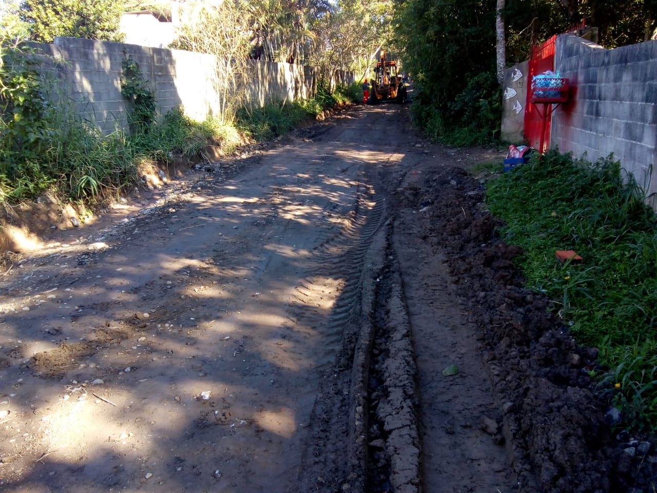 #PraCegoVer - O leito da rua é de terra, que está sendo regularizado. Ao fundo há uma máquina que vai nivelar a rua. À esquerda, há um muro. À direita, outro muro com um portão vermelho.