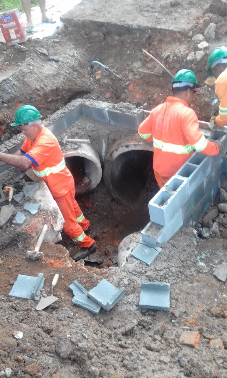 #PraCegoVer - Trabalhadores constroem caixa de escoamento. Em um buraco cavado no chão, apontam dois tubos grandes que chegam à caixa, feita de tijolos de cimento.
