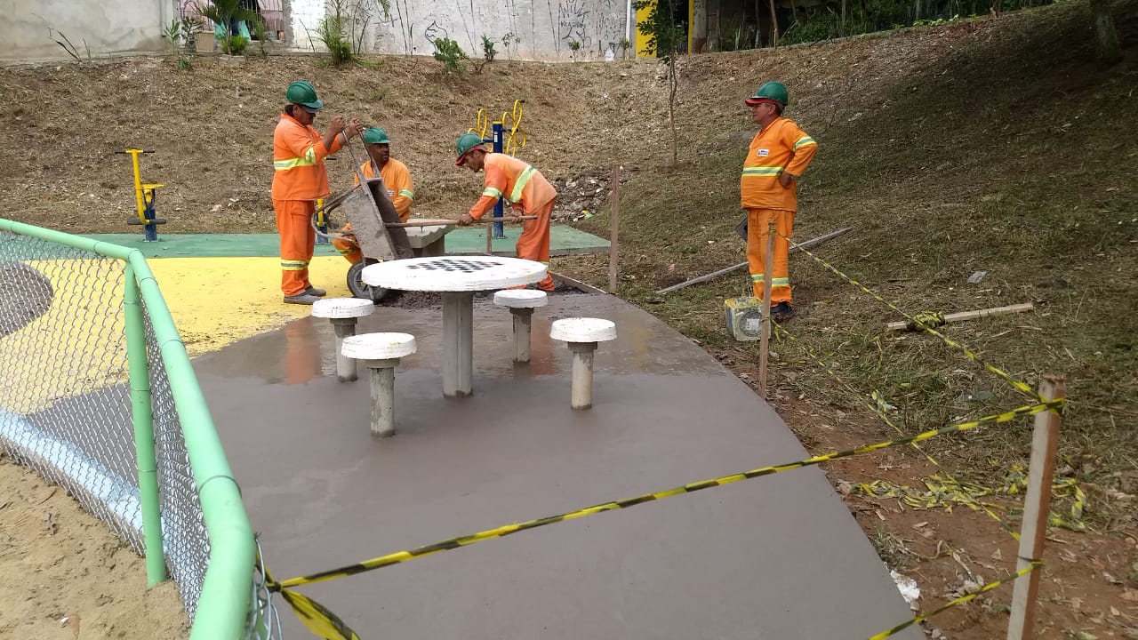 #PraCegoVer - Quatro trabalhadores da Subprefeitura, em macacões cor de laranja, finalizam o piso da   praça. No local, há uma mesa com tabuleiro de jogos, cercada por quatro bancos pintados de branco.