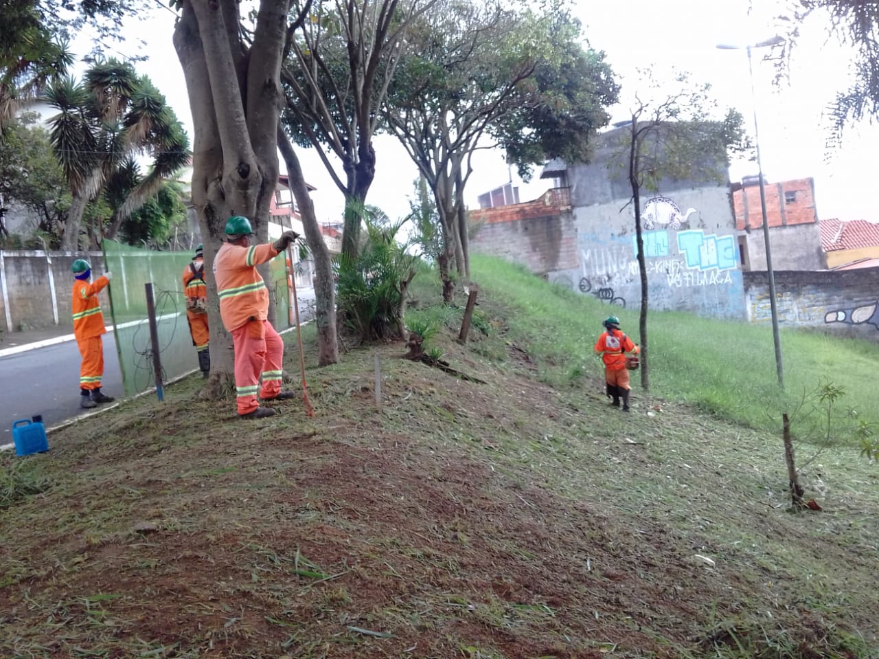 #PraCegoVer - Trabalhadores da Subprefeitura cortam a grama na praça. O corte está sendo feito em uma   encosta. Há árvores do lado esquerdo.