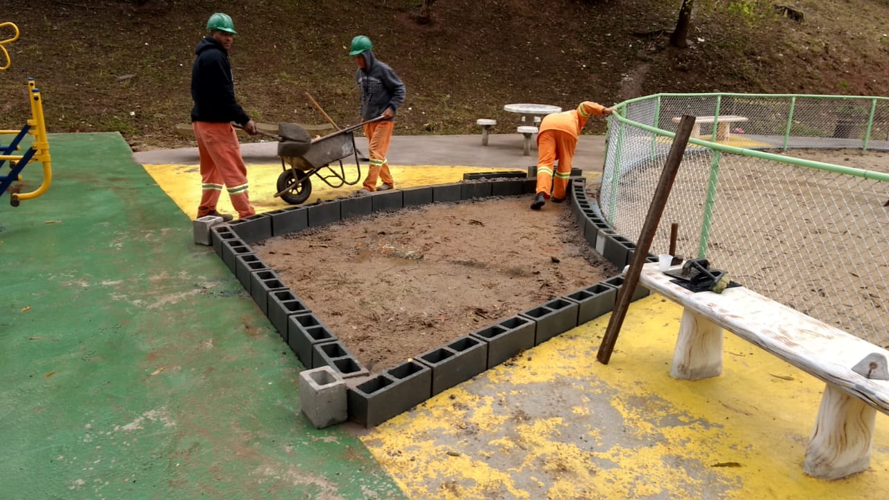 #PraCegoVer - Trabalhadores da Subprefeitura constroem um cercado de tijolos perto da caixa de areia. Um dos trabalhadores assenta os blocos. Outro maneja um carrinho de mão.