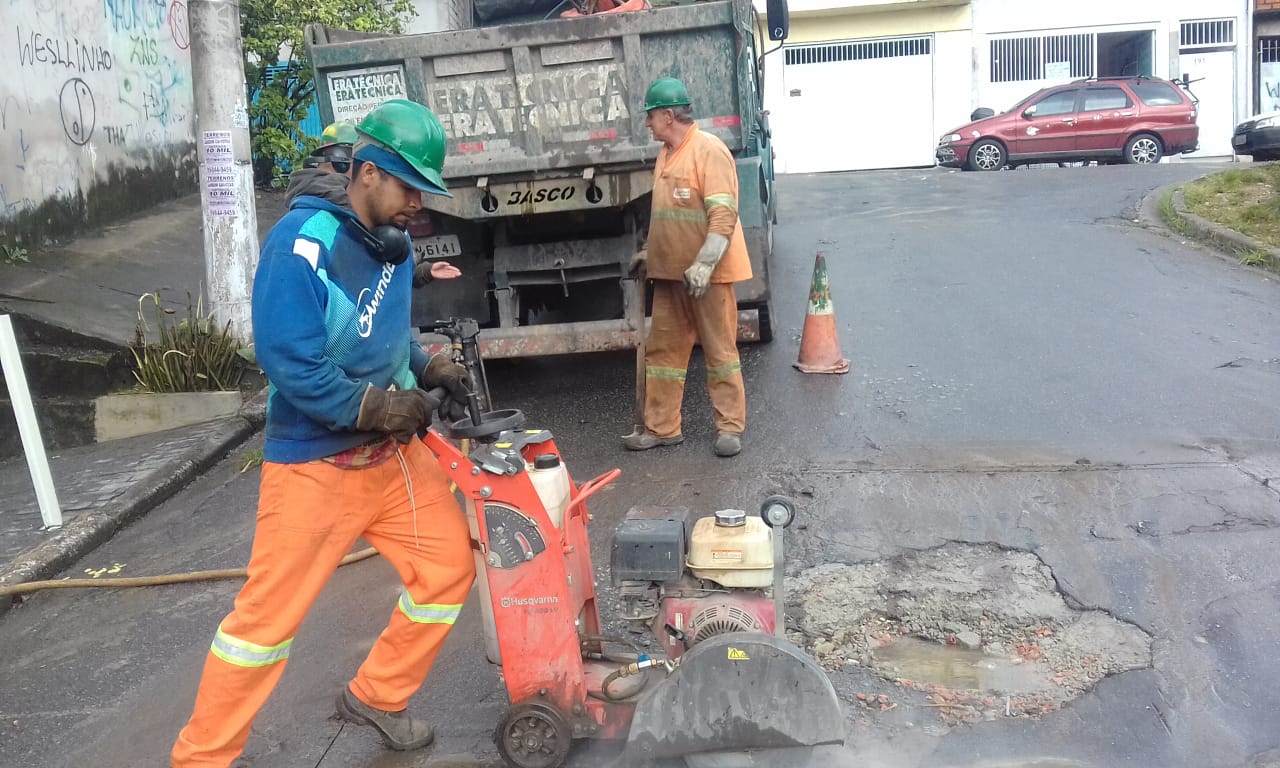 #PraCegoVer - Trabalhador de Subprefeitura usa máquina para recortar o asfalto em volta do buraco na via. Ao fundo, outro trabalhador junto ao caminhão da empreiteira contratada.