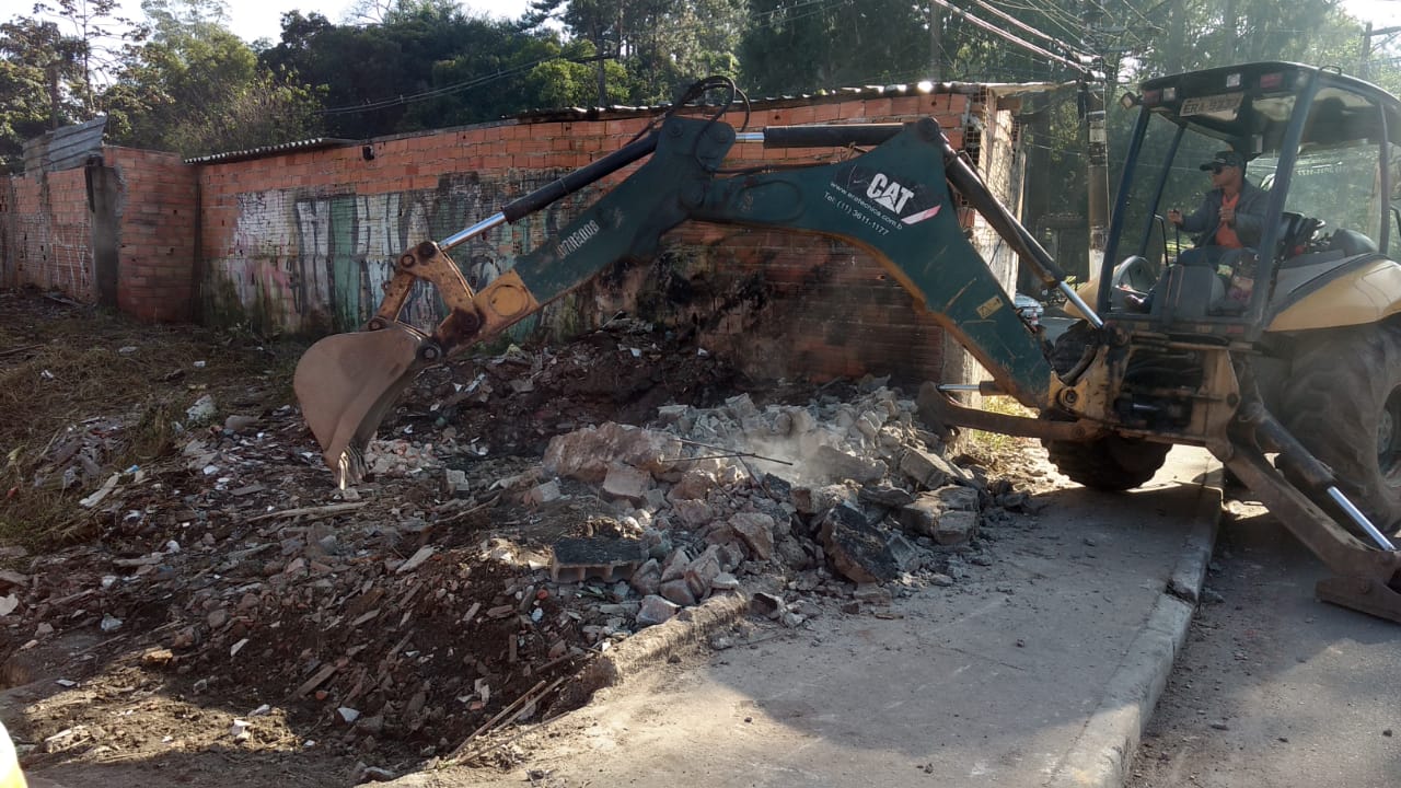 #PraCegoVer - Uma escavadeira verde, apoiada na calçada, limpa entulho produzido por muro derrubado. 