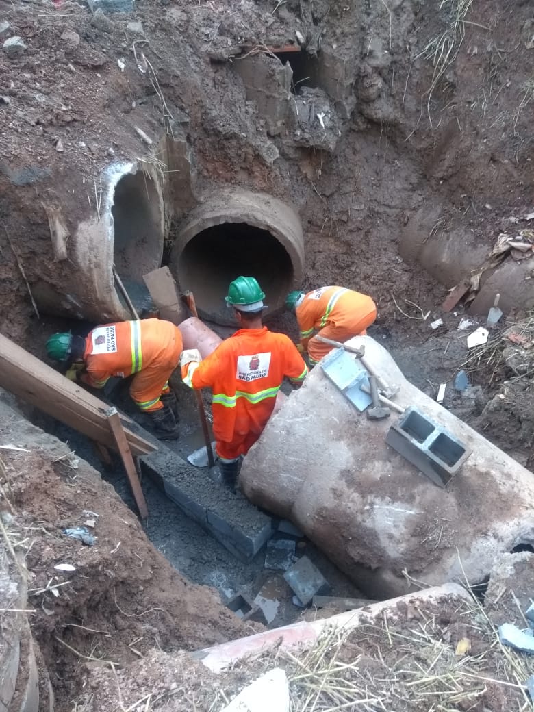 #PraCegoVer - Trabalhadores da Subprefeitura fazem a ligação de tubos de escoamento no fundo de um buraco cavado no local.