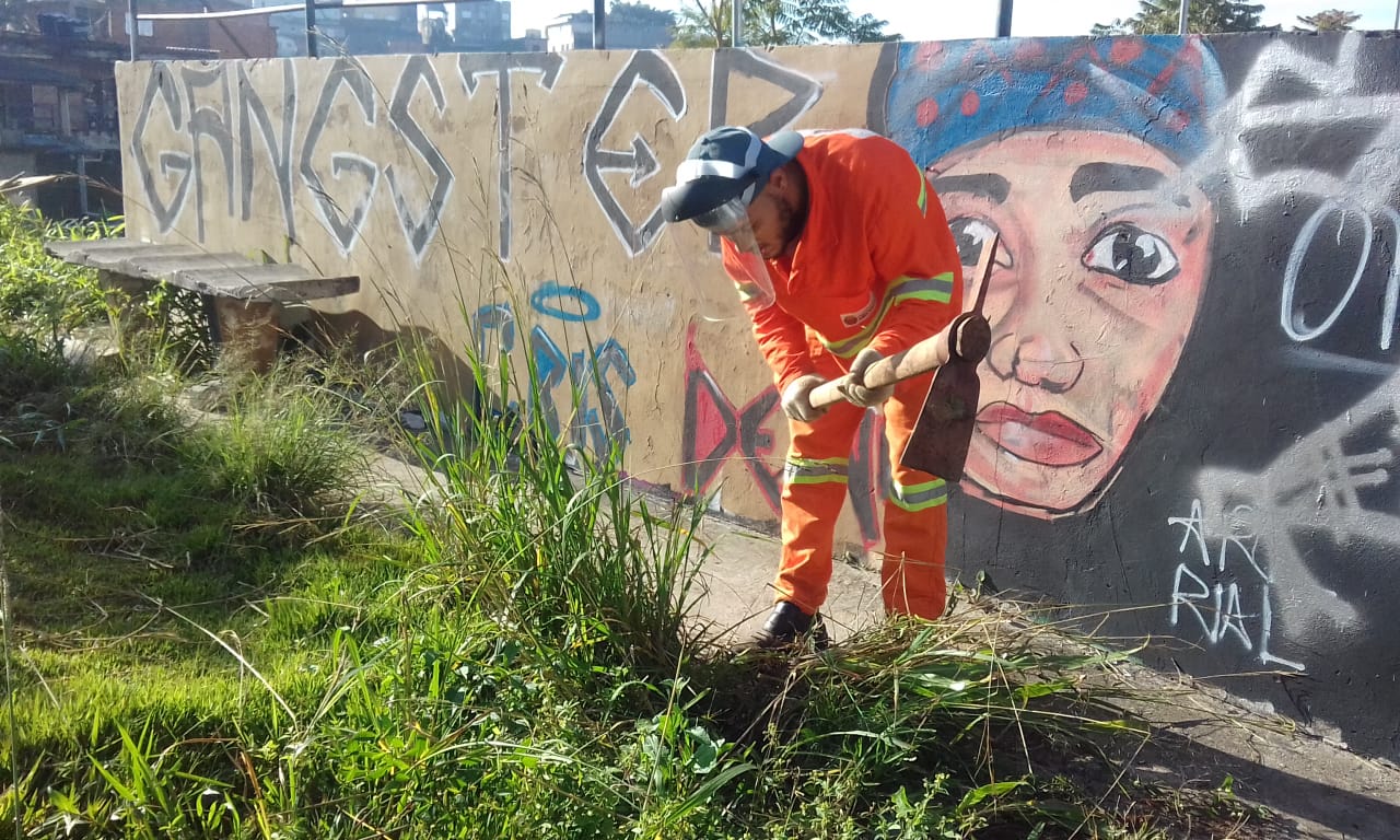 #PraCegoVer - Um trabalhador da Subprefeitura usa uma picareta para arrancar mato do local. O trabalhador usa uma viseira de proteção. Ao fundo, há um muro com um grafite de um rosto e a palavra "Gangster" ao lado.