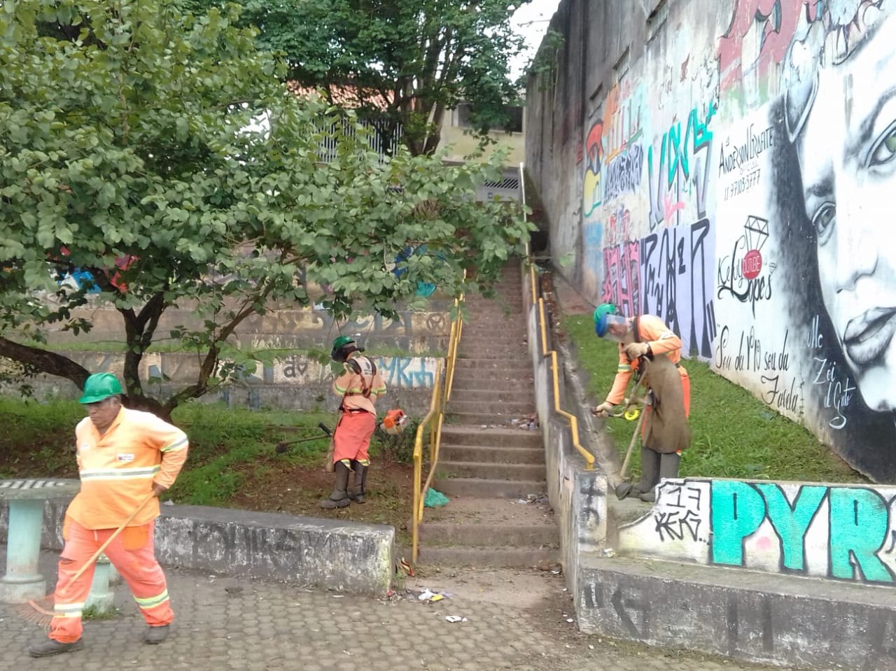 #PraCegoVer - Trabalhadores cortam grama e recolhem grama e folhas em uma escadaria ladeada por   corrimãos amarelos. A escadaria fica ao lado de uma parede em que há vários grafites.