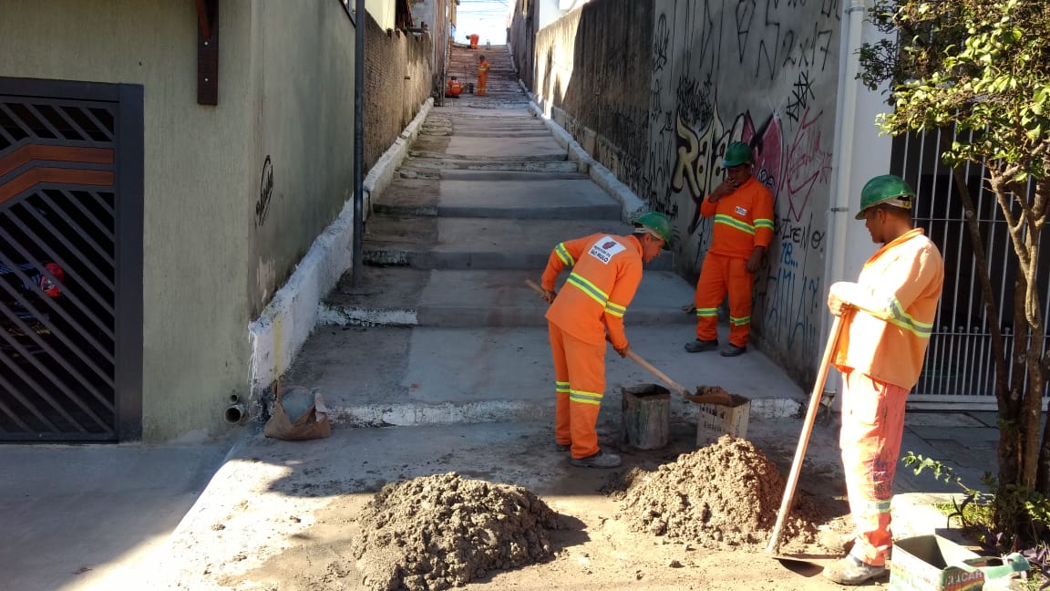 #PraCegoVer - Três trabalhadores da Subprefeitura em primeiro plano, misturam cimento e areia para a massa que vai cobrir os degraus do escadão. Outra turma, de mais três trabalhadores, estão no alto do escadão.