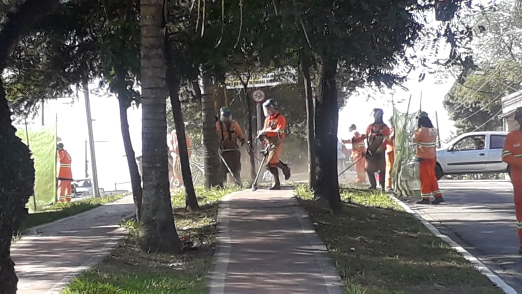 #PraCegoVer - Nove trabalhadores da Subprefeitura cortam grama no canteiro central da avenida Teotônio   Vilela, entre as pistas da ciclovia. Três deles manejam os cortadores portáteis. Os demais protegem o   local com telas. Outros, ainda, varrem o produto do corte da grama.