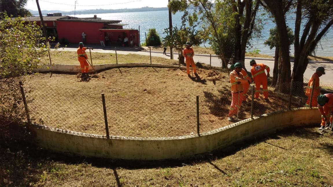 #PraCegoVer - Sete trabalhadores da Subprefeitura limpam área cercada que será destinada a abrigar   brinquedos para crianças. Ao fundo, aparece a represa Billings.