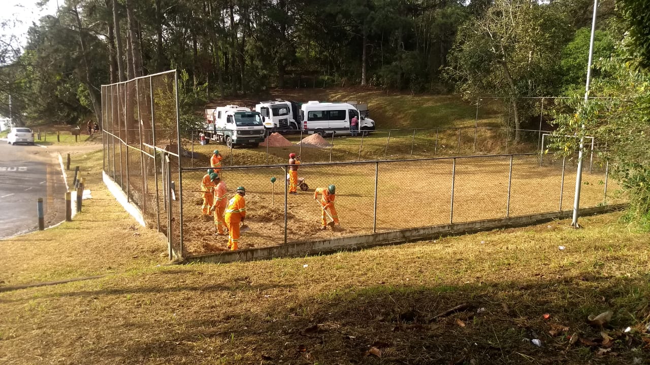 #PraCegoVer - Seis trabalhadores da Subprefeiura, com enxadas, limpam a superfície de uma quadra de   grama. A quadra é cercada por alambrado. Ao fundo, estão vans da Subprefeitura.