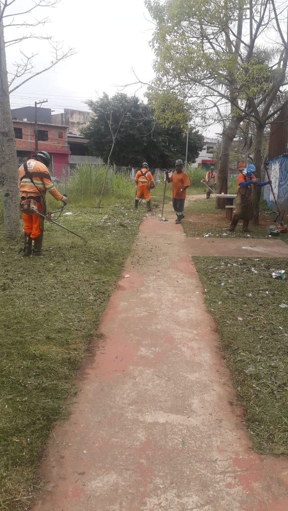 #PraCegoVer - Seis trabalhadores da Subprefeitura cortam grama adjacente a uma calçada interna. Há duas   árvores do lado direito. E casas ao fundo.