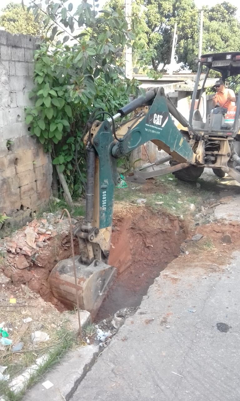 #PraCegoVer - Escavadeira verde, operada por trabalhador da Subprefeitura, abre uma vala onde será   instalado um novo coletor de boca-de-lobo. Há uma folhagem verde à esquerda, encostada a um muro.