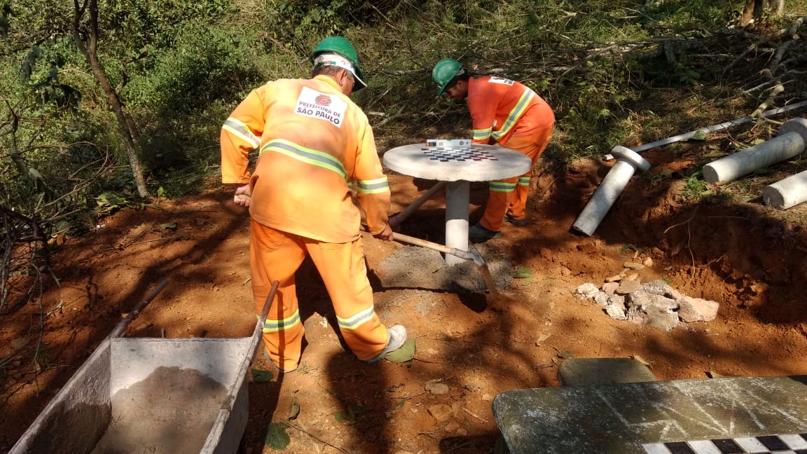 #PraCegoVer - Dois trabalhadores da Subprefeitura instalam mesas de concreto com tabuleiro de jogos e bancos de concreto.