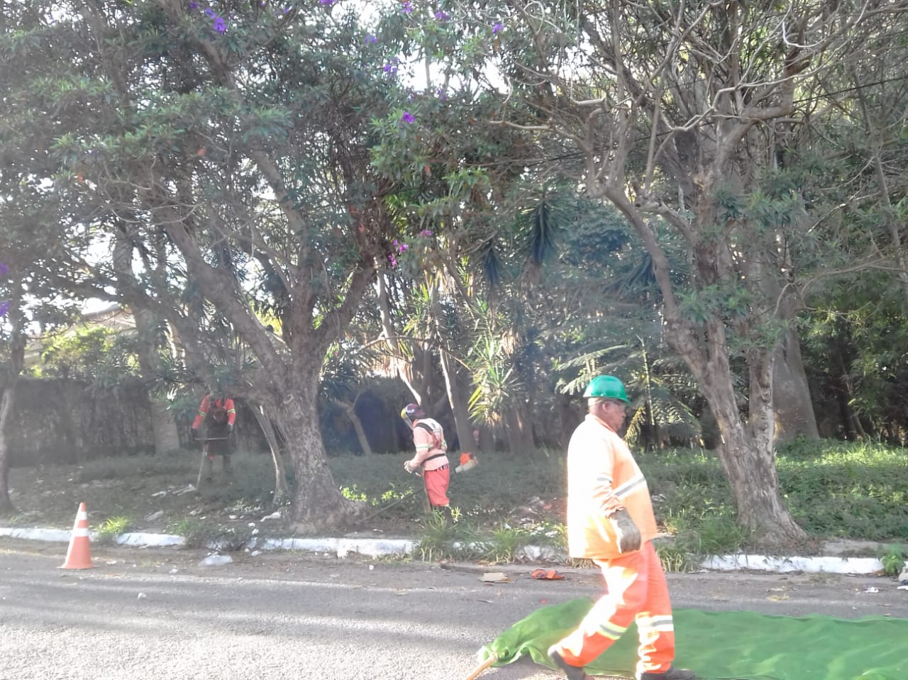 #PraCegoVer - Dois trabalhadores da Subprefeitura cortam grama. Um terceiro prepara-se para erguer a tela de proteção.