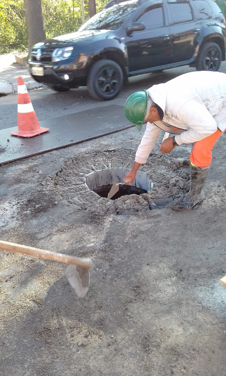 #PraCegoVer - Trabalhador da Subprefeitura faz o acabamento do poço-de-visita (PV), ao lado da via, sinalizada por um cone branco e laranja. Ao fundo passa um carro preto.